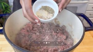 Adding spices to meat in a pot for a savory cooking recipe, with onions and herbs visible.