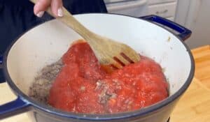 Stirring tomato sauce into ground beef in a blue Dutch oven pot on a wooden counter for a homemade pasta dish.