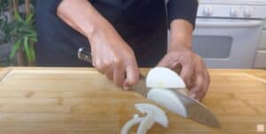 Person slicing an onion on a wooden cutting board in a kitchen setting.