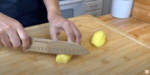 Person slicing a potato with a chef's knife on a wooden cutting board in a kitchen.