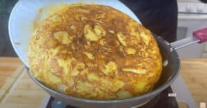 Golden Spanish tortilla being flipped from a frying pan onto a plate in a kitchen setting.