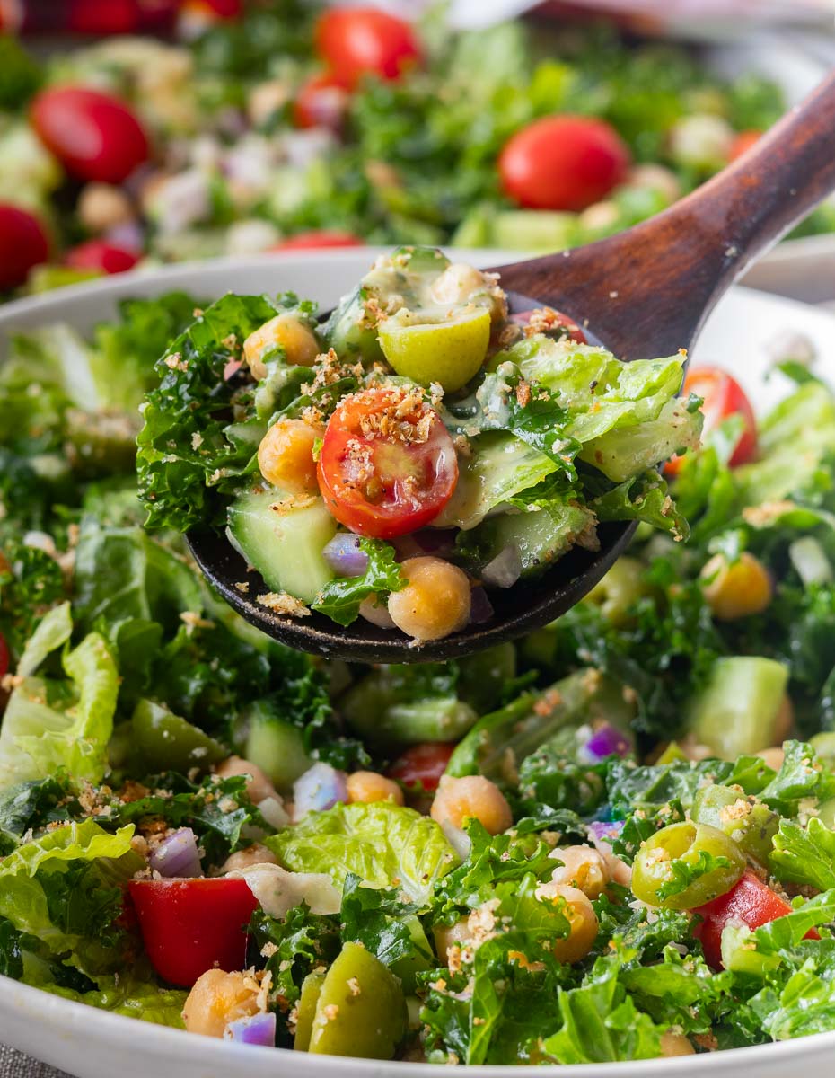 Hummus Crunch Salad with kale, cherry tomatoes, chickpeas, onions, and cucumbers in a wooden spoon.