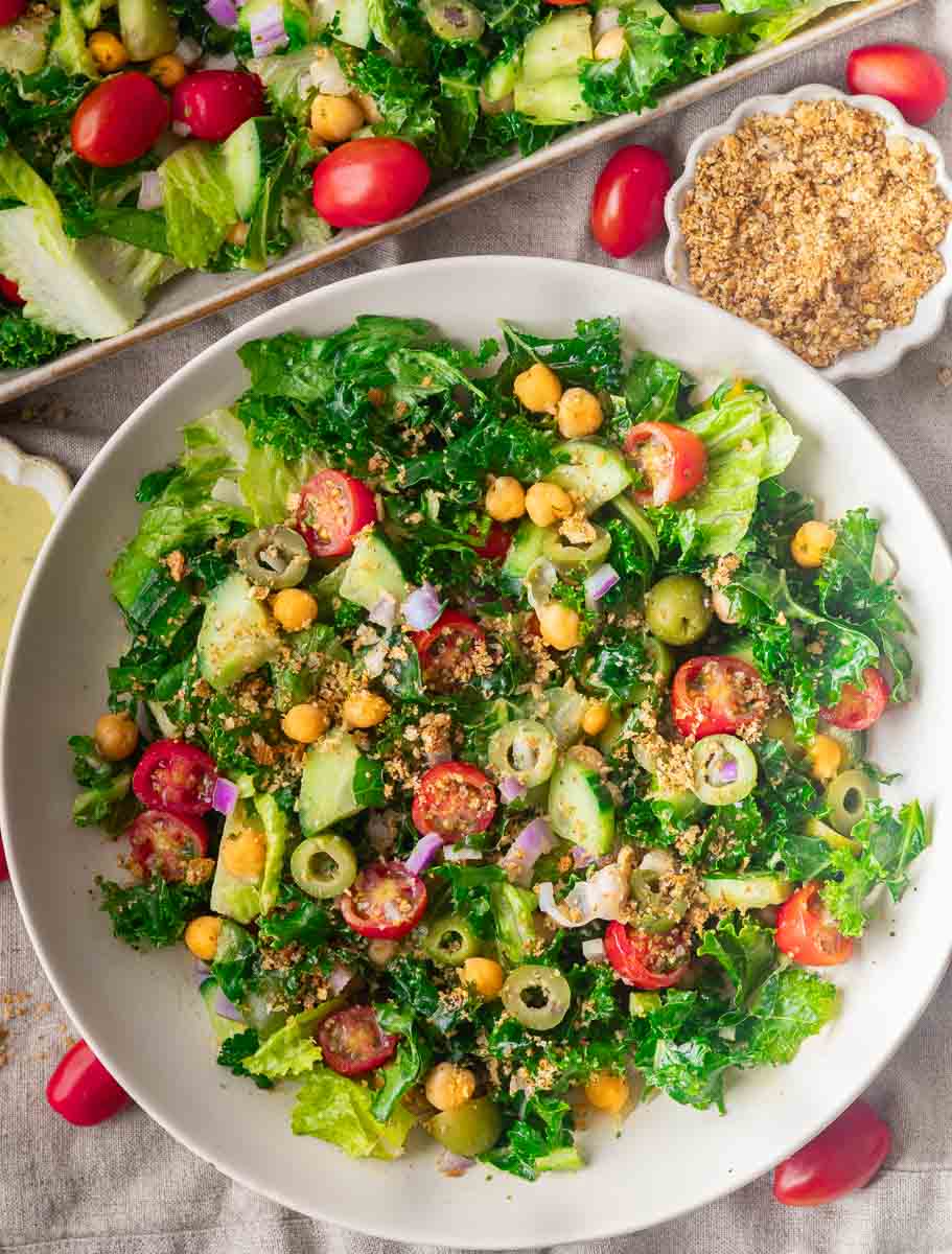 Hummus Crunch Salad with kale, cherry tomatoes, olives, cucumbers, and chickpeas in a white bowl, garnished with breadcrumbs.
