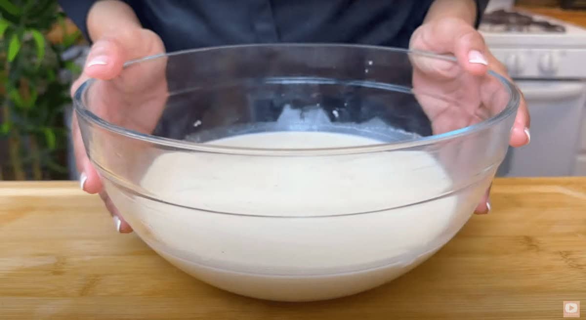 Person holding a clear glass bowl of crepe batter on a wooden countertop.