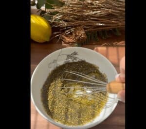 Whisk mixing spices in a bowl beside a lemon on wooden surface with herbs in background.