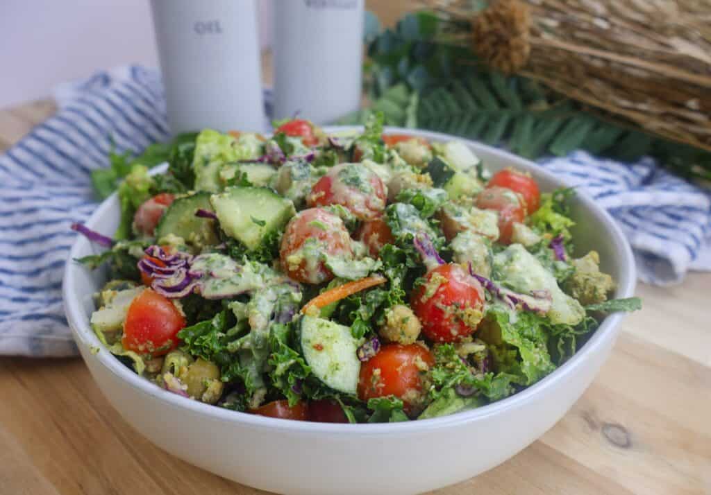 Fresh garden salad with cherry tomatoes, cucumbers, kale, and creamy dressing in a white bowl on a wooden table.