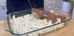 Layering meat sauce over creamy pasta in a glass baking dish on a wooden countertop.