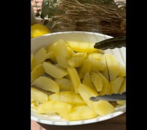 Sliced seasoned potatoes in a white bowl with black tongs, brightened by sunlight, ready for cooking.