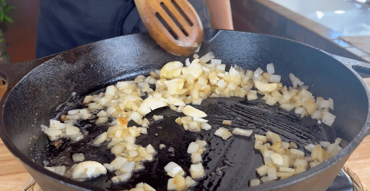 Sautéing chopped onions and garlic in a cast iron skillet, cooking in progress with a wooden spatula.