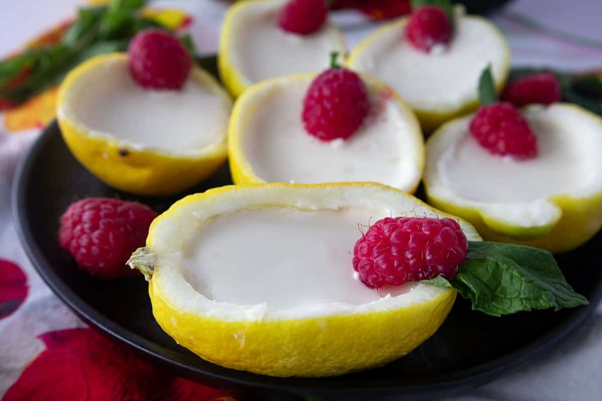 Lemon cups filled with creamy dessert, topped with fresh raspberries and mint leaves, served on a black plate.