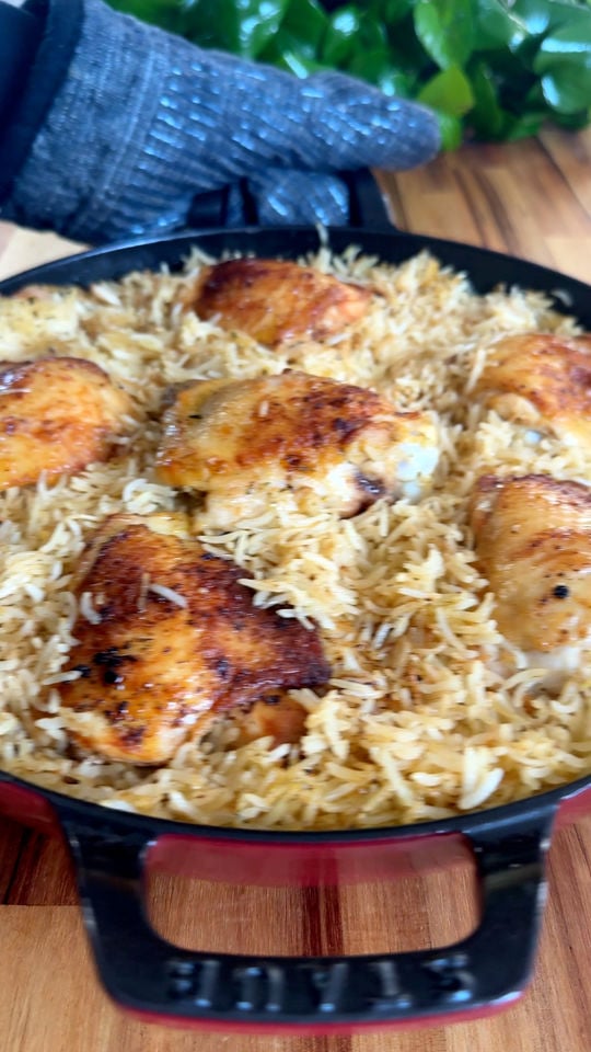 Cast iron pan with baked chicken thighs and rice, garnished with spices, on a wooden board with a gloved hand.