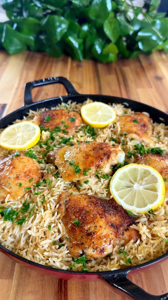Skillet of seasoned chicken thighs with rice, topped with lemon slices and parsley, on a wooden table.