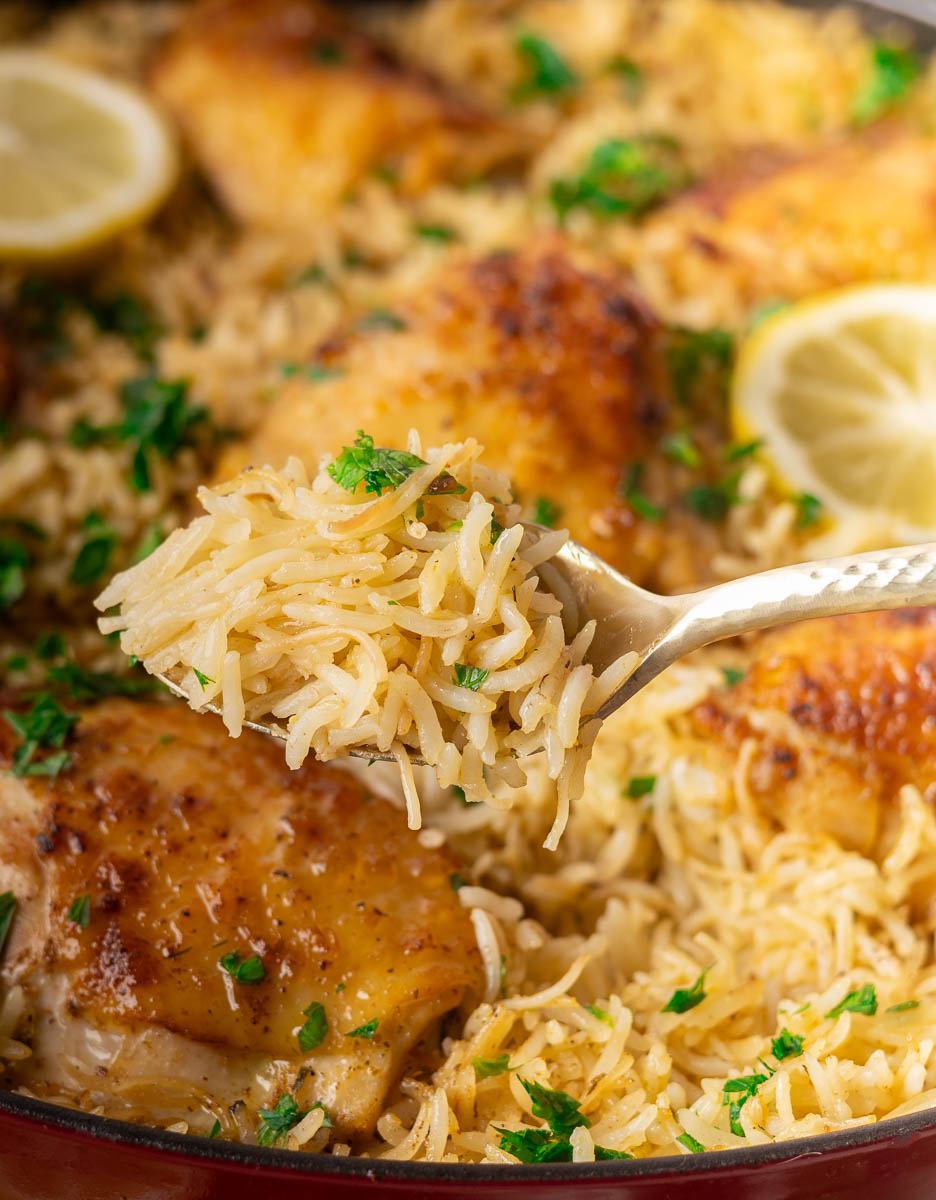 Spoon holding cooked rice with parsley over roasted chicken and lemon slices in a pan.