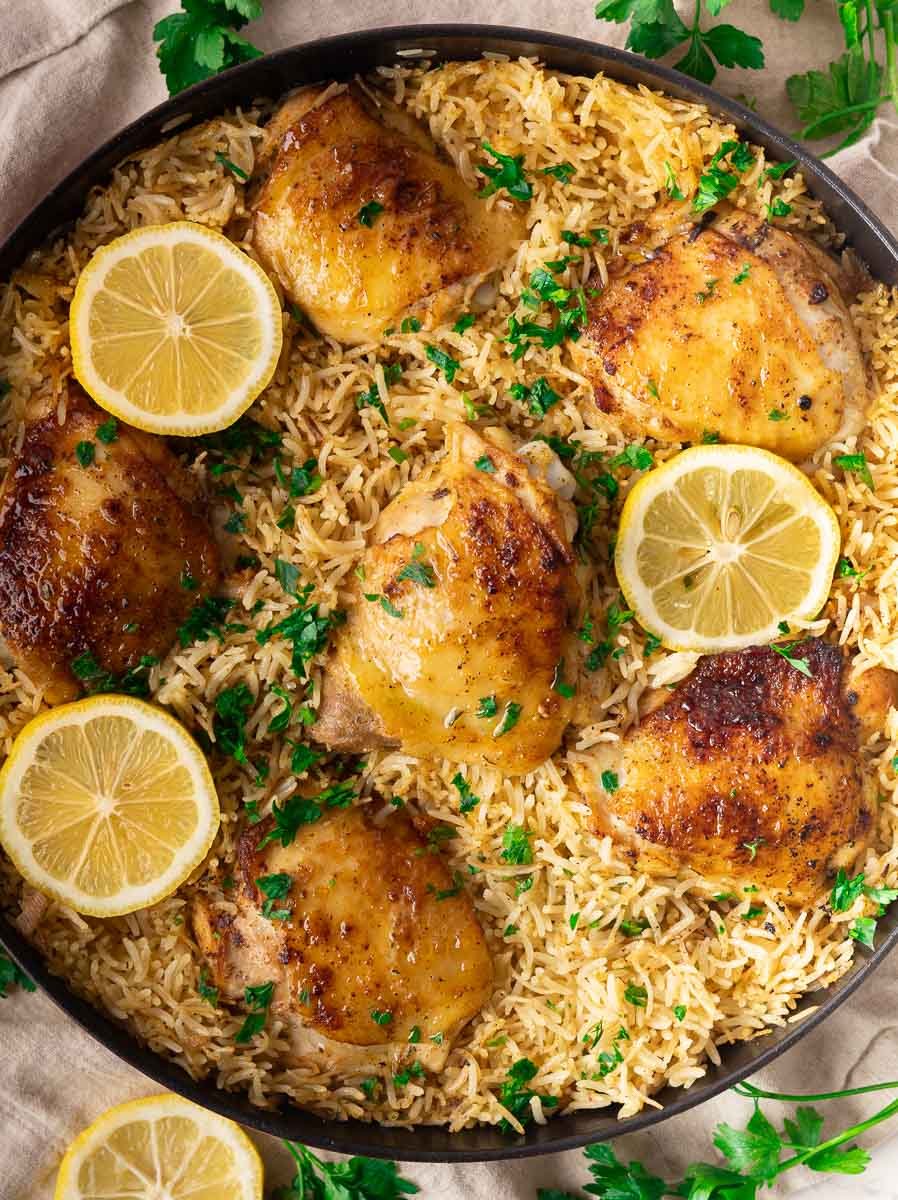 Lemon garlic chicken and rice pilaf with seasoned rice with lemon slices and parsley garnish in a black pan.
