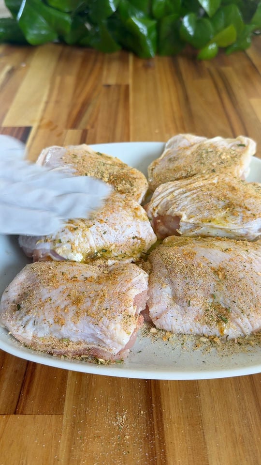 Raw chicken thighs seasoned with spices on a white plate, gloved hand adjusting them on wooden table.