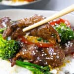 Beef stir-fry with broccoli, red peppers, and sesame seeds on rice, held with chopsticks. Asian cuisine dish.