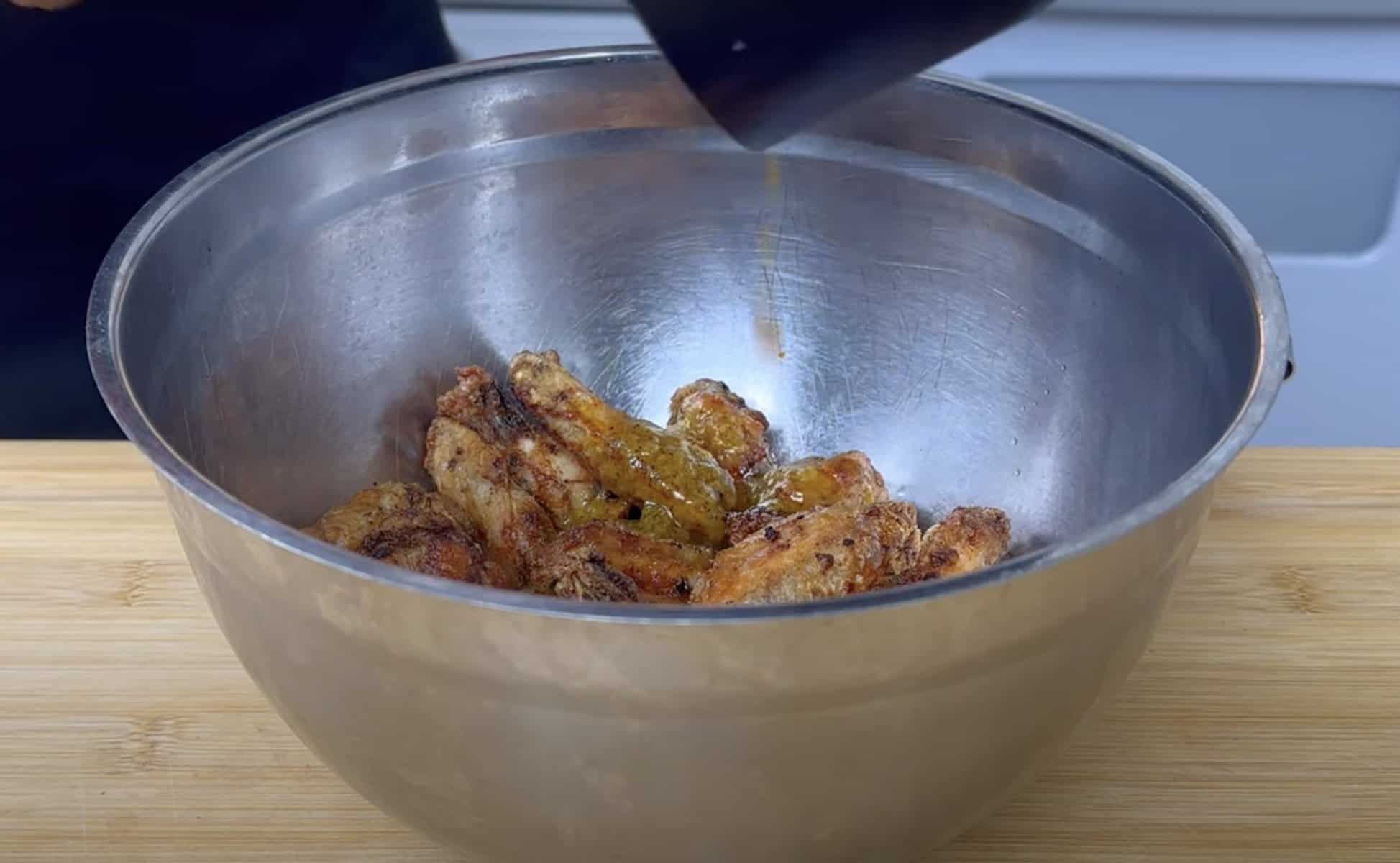 Pouring sauce over crispy chicken wings in a stainless steel bowl on a wooden surface.