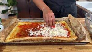 Hand sprinkling cheese on a homemade pizza with tomato sauce on parchment-lined baking sheet.