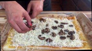 Hands adding mushrooms to cheesy puff pastry on parchment paper.