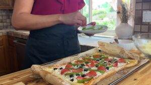 Woman preparing homemade pizza with bell peppers and olives in a bright kitchen.