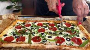 Person cutting a fresh homemade pizza with pepperoni, olives, and green peppers using red scissors.