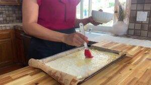 Person brushing olive oil on filo pastry sheet in a kitchen. Baking preparation on wooden countertop with natural light.