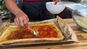 Person spreading tomato sauce on puff pastry for homemade pizza preparation.