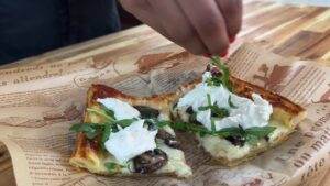 Person garnishing gourmet mushroom tart with arugula and creamy cheese on rustic paper background.