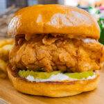 Crispy fried chicken sandwich with pickles on a brioche bun, served with fries on a wooden plate.
