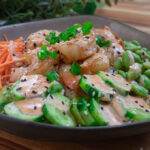 Colorful shrimp poke bowl with carrots, edamame, cucumbers, and spicy mayo dressing on a wooden table.