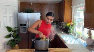 Woman in kitchen cooking spaghetti in pot, surrounded by wooden cabinets and flowers by the window.