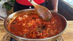 Person cooking rich tomato sauce with chunks of fish in a skillet, stirring with a wooden spoon.