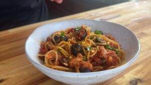 Delicious spaghetti with tomato sauce, olives, and herbs in a white bowl on a wooden table. Perfect Italian meal.