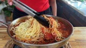 Person stirring spaghetti in tomato sauce with tongs in a kitchen setting.