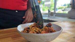 Person serving delicious spaghetti with tongs in a white bowl, set on a wooden kitchen counter.