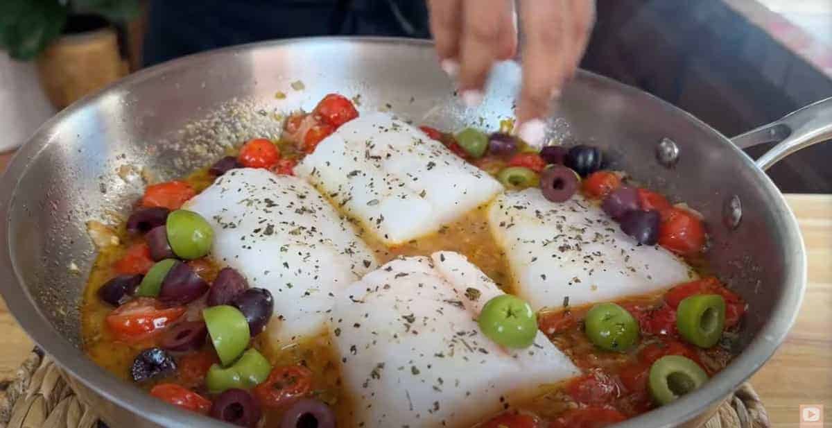 Cooking cod in a skillet with cherry tomatoes and olives.