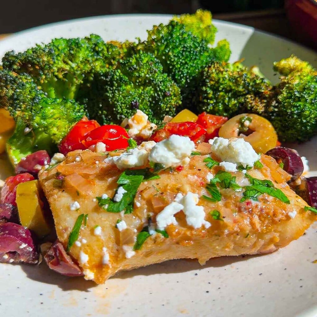 Mediterranean cod with feta, olives, tomatoes, and parsley, served with roasted broccoli on a white plate.