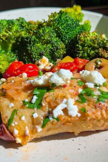 Mediterranean cod with feta, olives, tomatoes, and parsley, served with roasted broccoli on a white plate.