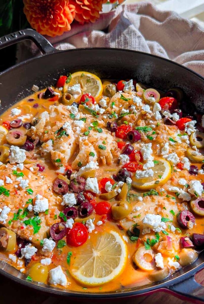 Skillet with Mediterranean cod, olives, feta cheese, lemon slices, and herbs in a flavorful sauce.