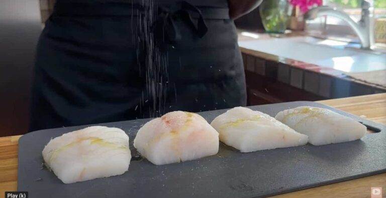 Person seasoning four pieces of raw fish on a cutting board in a kitchen setting.