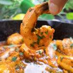 Close-up of hand holding a spicy shrimp topped with fresh herbs, cooked in flavorful sauce in a skillet.