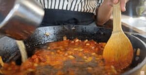 Cooking tomato sauce in a cast-iron skillet with a wooden spoon and pouring broth for a simmering recipe.