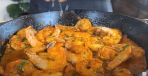 Sizzling shrimp in garlic tomato sauce with herbs in a cast iron skillet, a flavorful seafood delicacy being garnished.