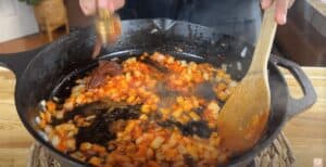 Cooking onions and spices in a cast iron skillet with a wooden spoon, adding seasoning, in a cozy kitchen setting.