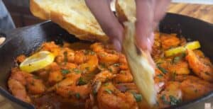 Spicy shrimp in a skillet with lemon slices and bread, garnished with herbs for a flavorful seafood dish.