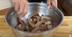 Person mixing shrimp with spices in a stainless steel bowl on a wooden countertop. Perfect for seafood recipes.