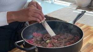 Grating garlic into a pan of sizzling beef strips on a wooden kitchen countertop.