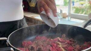 Person seasoning sizzling beef in a pan by a sunlit kitchen window.