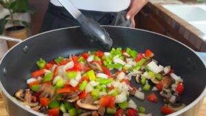 Sautéing colorful bell peppers, onions, and mushrooms in a black skillet with tongs. Fresh vegetables cooking.