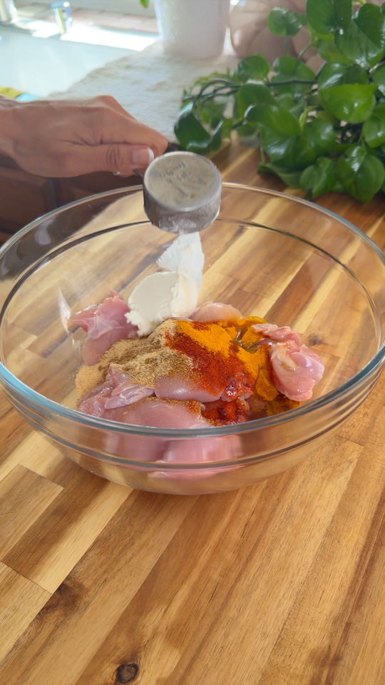 Person adding yogurt to bowl with chicken and spices on wooden surface.
